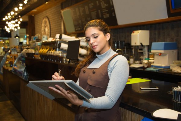 Portrait of confident young female cafe owner wearing uniform writing in register book or menu software Software confident cafe owner writing register book scaled 600x400