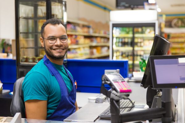 Smiling African American cashier sitting at checkout. Cheerful bearded young man in eyeglasses at workplace. Shopping concept software Software smiling african american cashier sitting checkout scaled 600x400